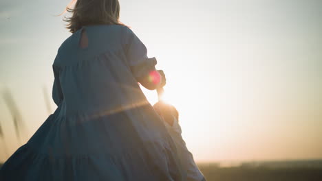 a joyful close-up captures a mother in a flowing blue gown spinning her little boy, dressed in a white top, in a grassy field during sunset. both are laughing