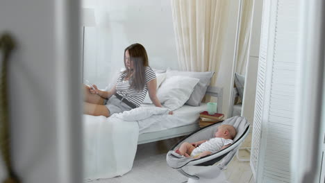 mom looks into the mobile phone, and next to the baby cradles a chair in a white interior.