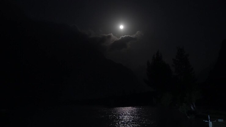 La luna piena brilla sopra un lago di montagna scuro, proiettando un riflesso luminoso sull'acqua.