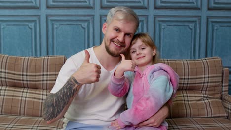 Father-and-little-child-daughter-kid-in-pajamas-sit-on-couch-in-room-smiling,-showing-thumbs-up