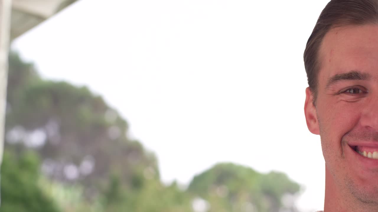Uomo adulto che sorride, con un sorriso ampio e gli occhi socchiusi, rilassandosi mentre viene ripreso da una telecamera vicino a un portico con alberi, spazio per il testo.