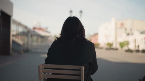 rear view of woman seated outdoors on wooden chair in urban setting, wearing black coat with soft natural light, blurred background of modern buildings, and open sky