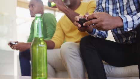 african american father and twin teenage sons sitting on couch playing game on tv and drinking beer