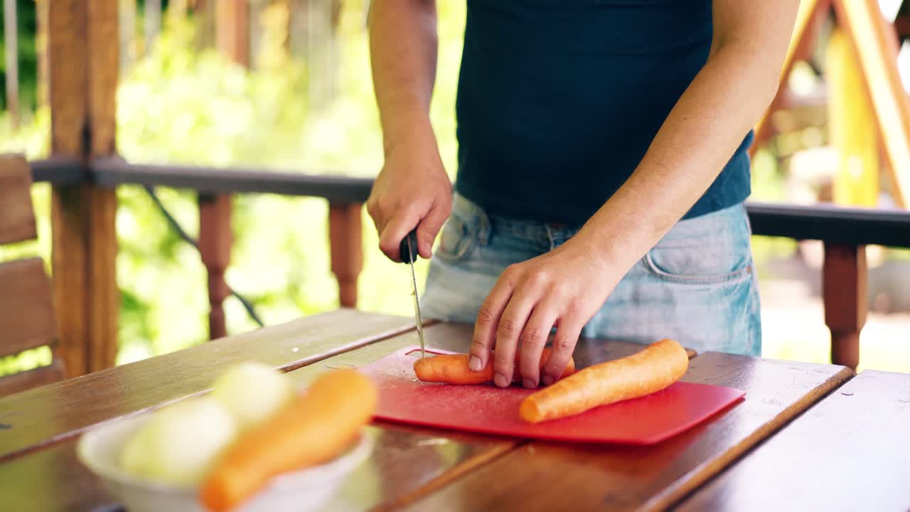 un joven está de pie en una casa de verano cocinando pilaf para un picnic. corta zanahorias en una tabla roja.