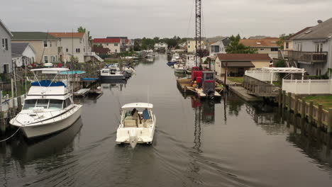 An-aerial-view-of-a-fishing-boat,-slowly-sailing-through-a-canal-behind-houses-on-Long-Island-on-a-cloudy-day