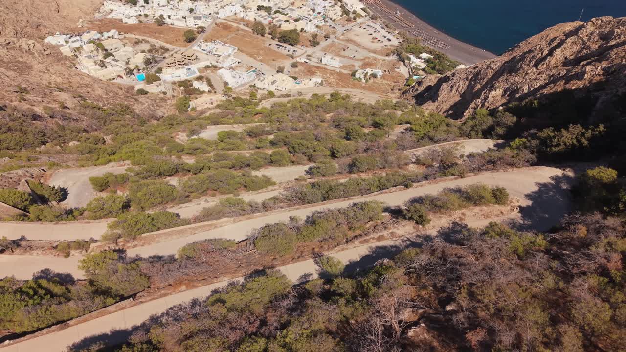 Una vasta veduta panoramica con drone che inizia sulle intense curve a gomito a zigzag che salgono sul monte Mesa Vouno, allontanandosi per rivelare l'intera estensione della spiaggia di Kamari e la scura costa vulcanica sottostante