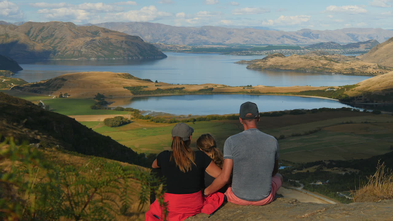 Family sit at hiking viewpoint by Lake Wanaka in New Zealand, from behind