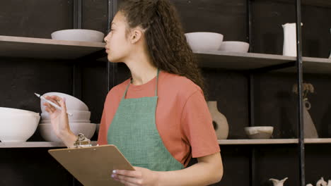 young woman writing on a clipboard and making an inventory of ceramics in the pottery shop