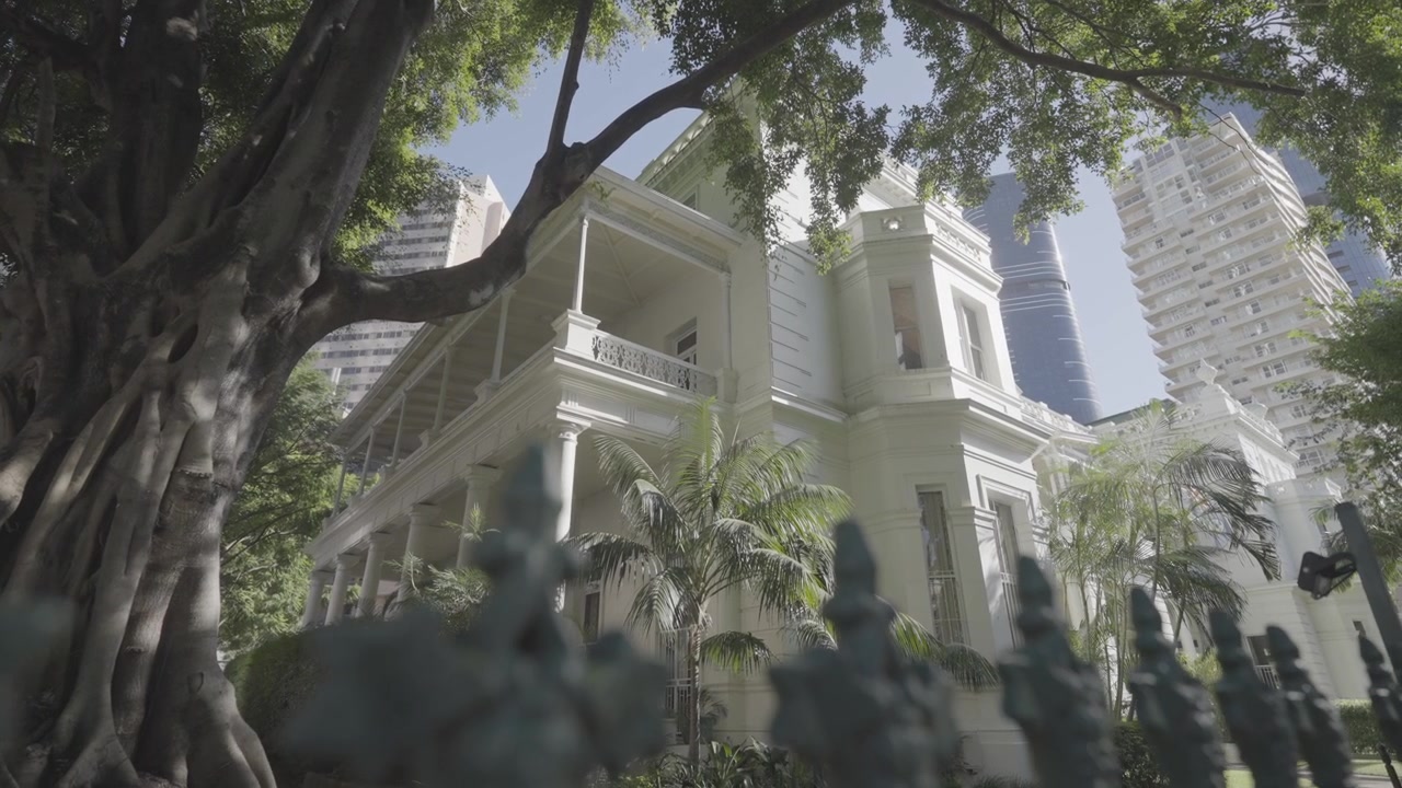 El balcón y las columnas blancas de un edificio histórico están enmarcados por palmeras, con modernas torres de la ciudad elevándose detrás