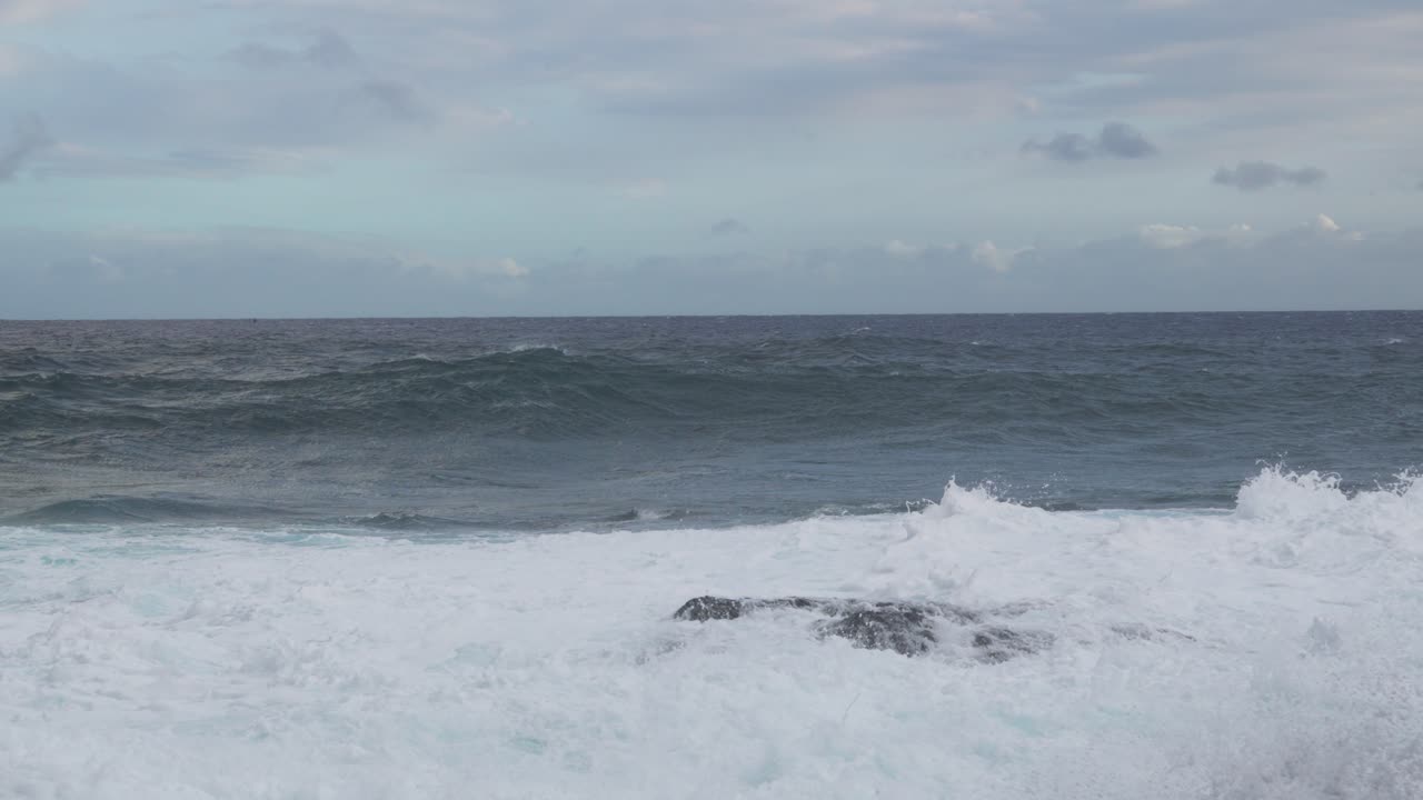 A windblown set rolls shoreward as a tall side plume lifts at frame left and foamy surge blankets the foreground, while a gray horizon sits beneath mottled, clouded sky