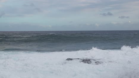 a windblown set rolls shoreward as a tall side plume lifts at frame left and foamy surge blankets the foreground, while a gray horizon sits beneath mottled, clouded sky