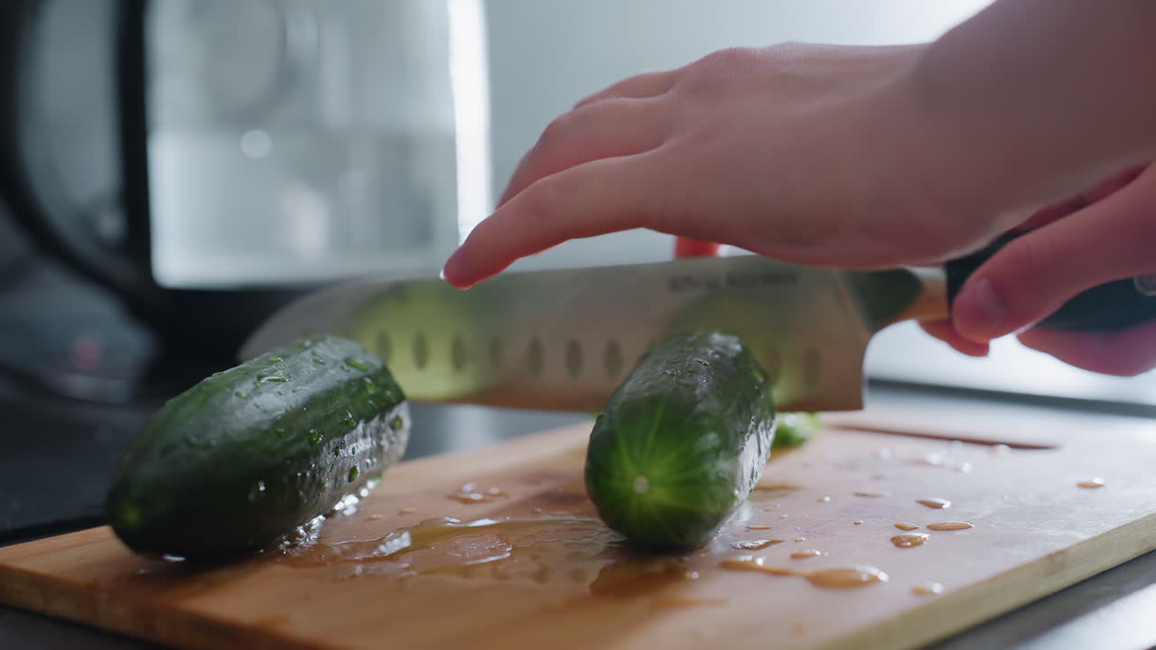 Close-up of hand slicing cucumber with sharp knife on wooden cutting board, kitchen scene with fresh ingredients, focusing on healthy food preparation and culinary action