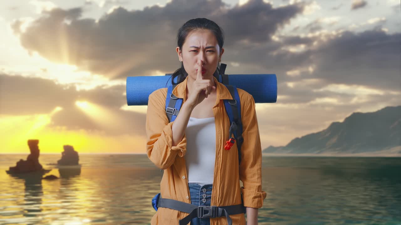 Pendaki Wanita Asia Dengan Ransel Gunung Melihat ke Arah Kamera dan Membuat Gestur Sst Sambil Berdiri di Pantai Saat Matahari Terbenam