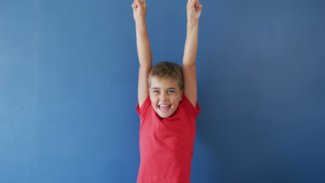 portrait of happy caucasian boy raising hands and smiling on blue background, slow motion