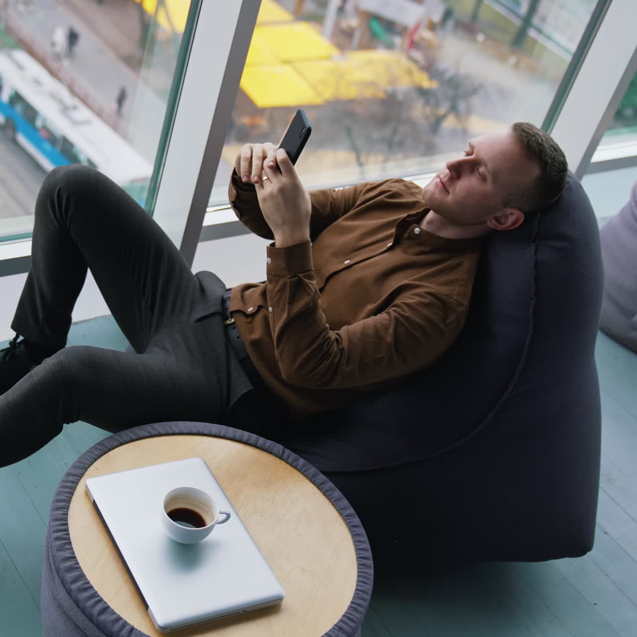 Man rests in a soft chair with a phone. Young man sitting near the window with a city transport background and using his mobile phone. Cup of coffee on small table. Top view