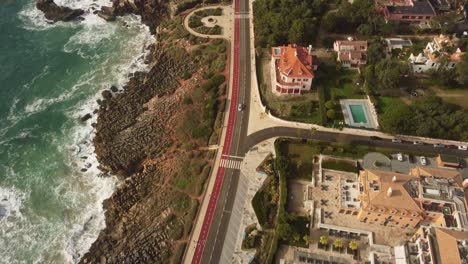 Vista-Aérea-De-La-Pequeña-Ciudad-De-Cascais-Portugal,-Pequeña-Carretera-Costera-Con-Coche-Conduciendo-A-Lo-Largo-Del-País-Cerca-De-La-Playa-Del-Acantilado-Rocoso-Del-Océano