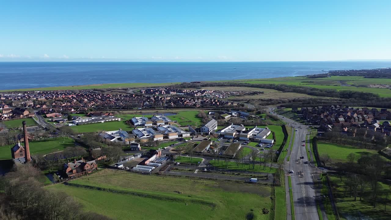 Sobrevoo de drone com movimento para frente sobre o campus do hospital psiquiátrico Hopewood Park, perto da costa galesa, mostrando estradas, campos, conjuntos habitacionais e o horizonte do mar.