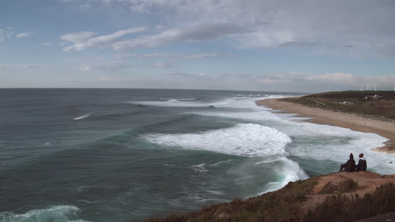 Two people sitting on a cliff overlooking a sandy beach watch the big waves rolling towards the shore