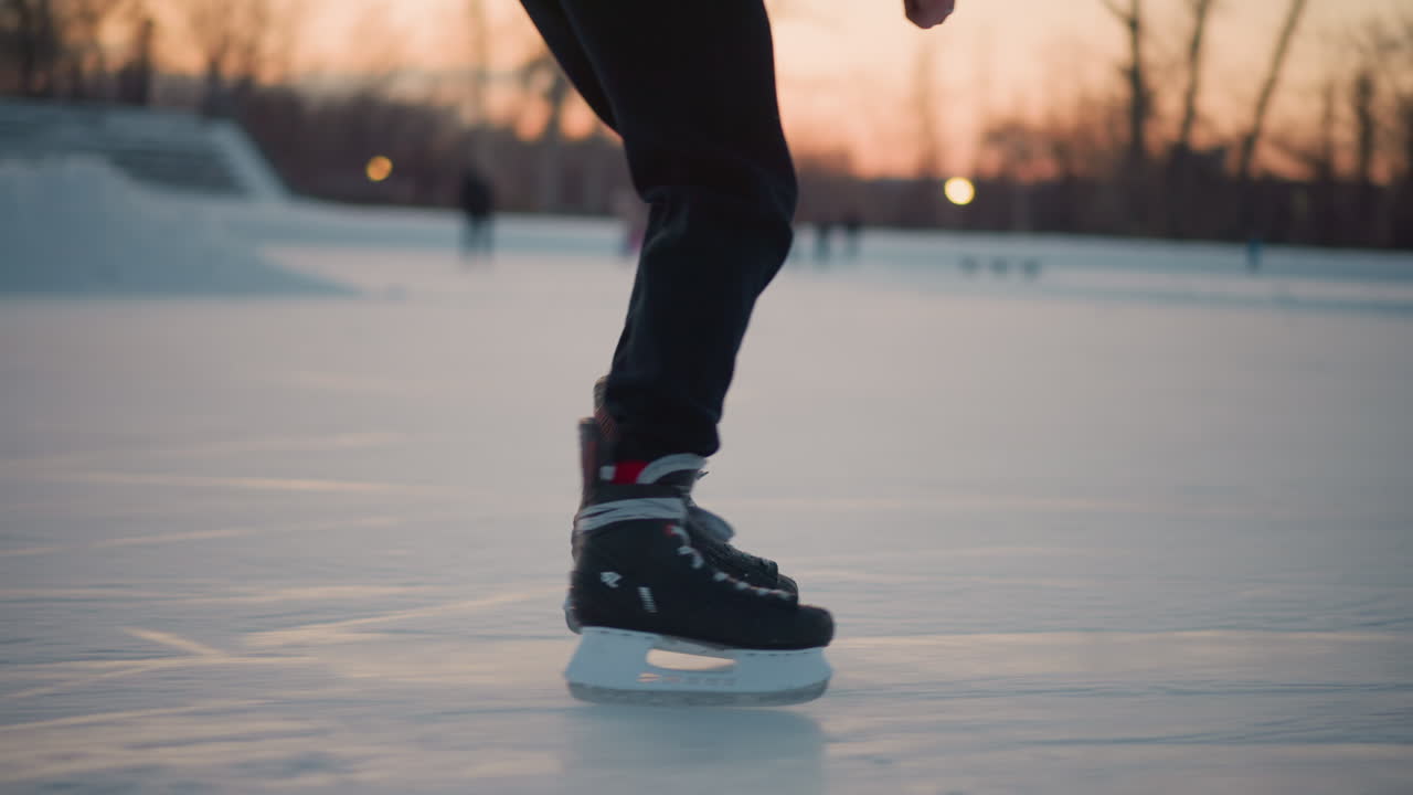 close up legs professional skater gliding gracefully on outdoor ice rink wearing black skates at sunset smooth movement in foreground with blurred figures of people skating in background