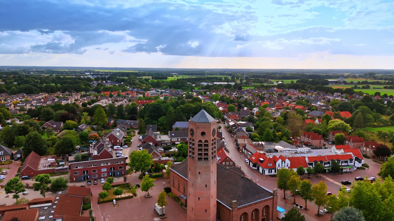 Vista aérea de una torre de iglesia de ladrillo y una plaza de pueblo europea. Vista de ángulo alto de una alta torre de iglesia de ladrillo en un barrio suburbano bajo un cielo nublado con rayos de sol y tejados rojos.
