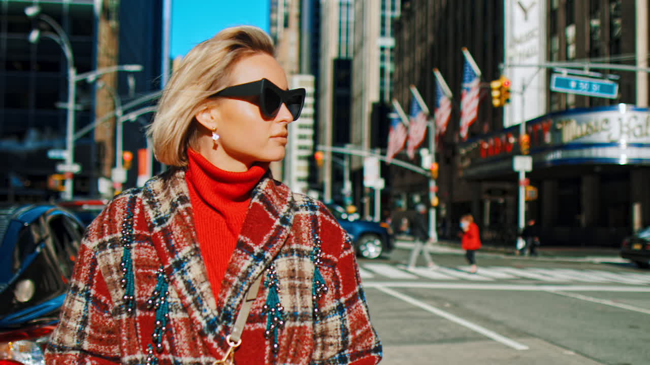New York, USA, 2 aprile 2026: Donna con occhiali da sole e cappotto a quadri a Midtown Manhattan. Donna bionda che indossa occhiali da sole e un cappotto di lana a quadri rossi su una strada di New York City vicino alla Music Hall.