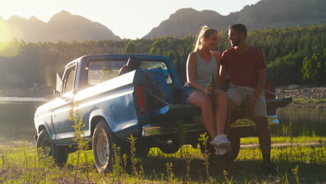 Couple-With-Backpacks-Sitting-On-Tailgate-Of-Pick-Up-Truck-On-Road-Trip-By-Lake-Drinking-Beer