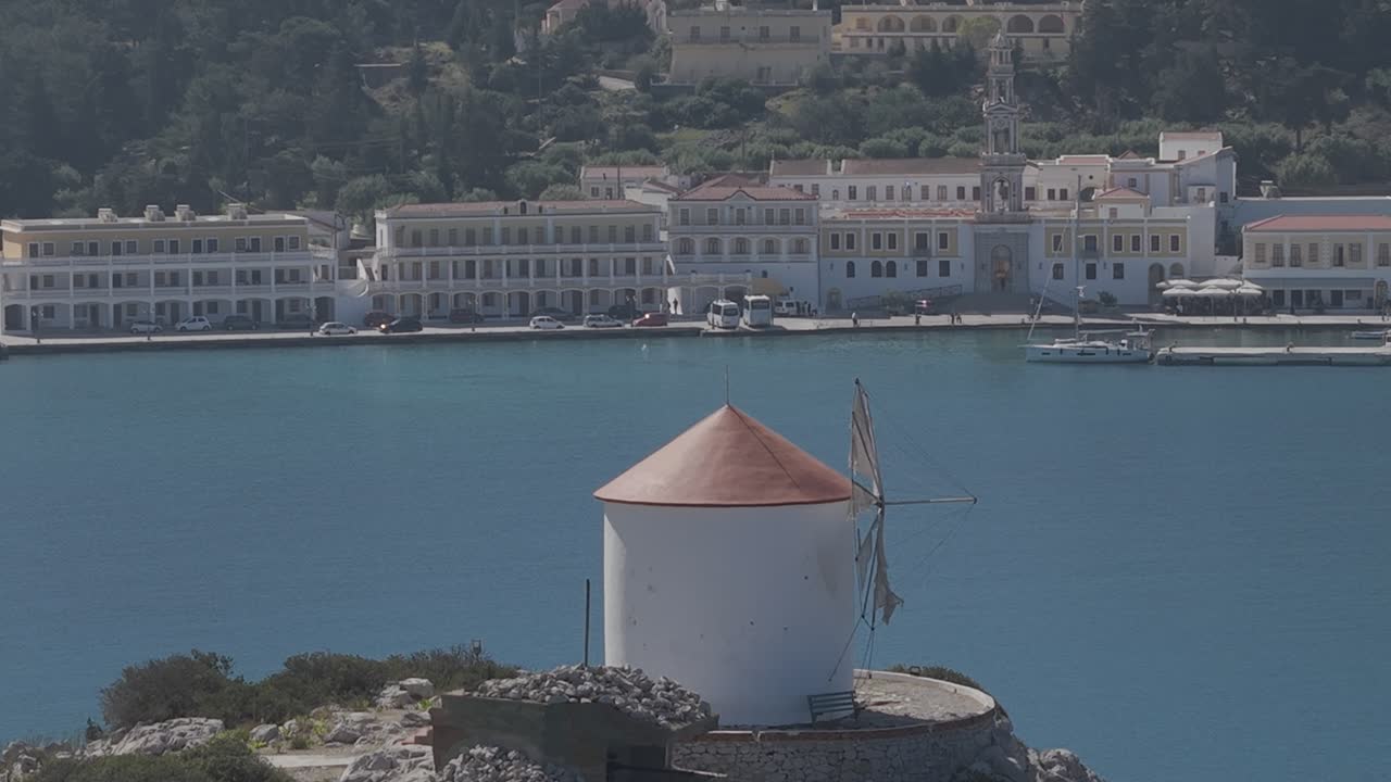 Stunning aerial perspective of the historic Panormitis Monastery on Symi. The footage showcases traditional Byzantine architecture in the background behind a windmill. 3x telephoto lens. D-Log M
