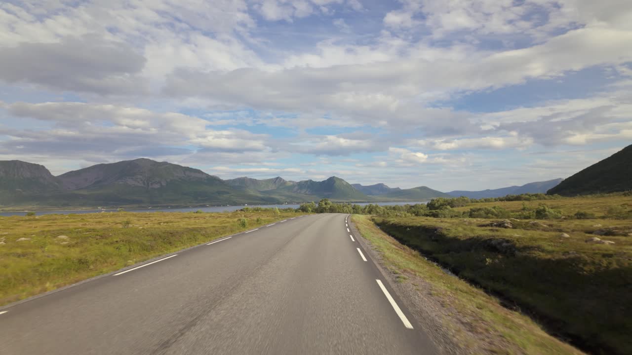 Andøya, Nordland, Noruega - Viagem por uma estrada local tranquila, ladeada por campos abertos e montanhas distantes sob um amplo céu nórdico - Plano em primeira pessoa
