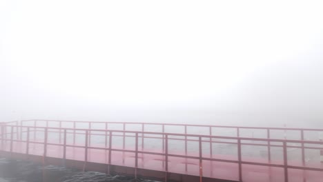 pan shot of red pier on calm lake disappearing into thick fog, slowly revealing misty shoreline and soft silhouettes in low contrast morning light in japan