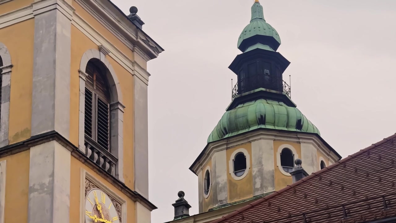 Primo piano della torre e della cupola verde della Cattedrale di Lubiana in Slovenia, che mostra un quadrante dell'orologio ornato, una facciata barocca gialla, un tetto in tegole rosse e un'architettura ecclesiastica storica.