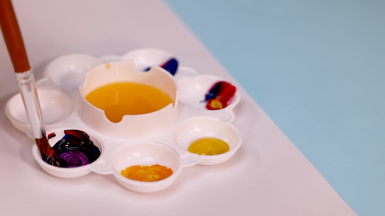 Brush stirs purple paint in white palette on pastel background, soft lighting, overhead view