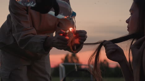 woman demonstrating fire control by pouring water from plastic bottle onto burning match held by girl against backdrop of twilight sky over grassy field