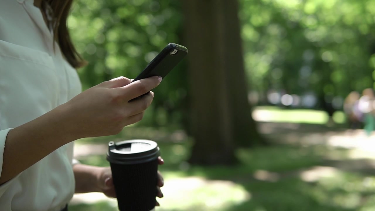 Donna che usa lo smartphone e tiene in mano una tazza di caffè al parco