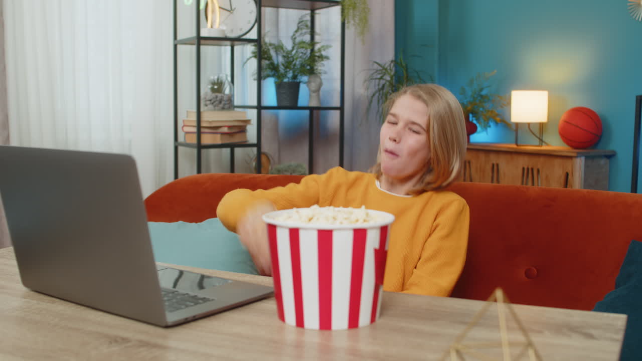 Teenager boy child watching funny video on laptop laughing while eating popcorn on couch at home