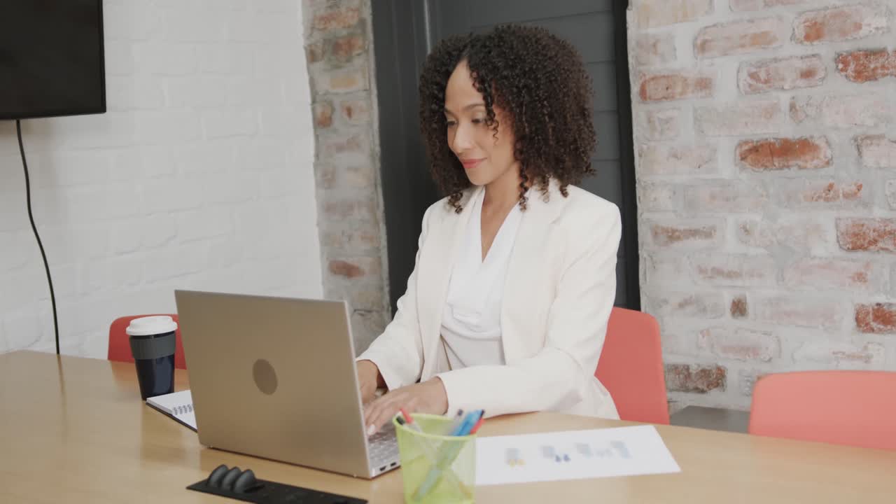Empresária birracial sorrindo sentada à mesa usando notebook no escritório, em câmera lenta.