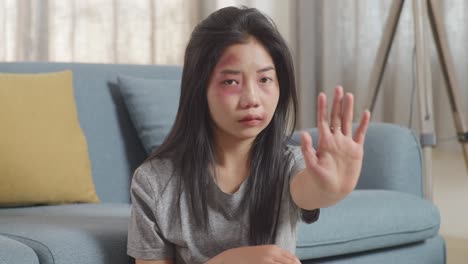 close up of young asian woman with bruise on body looking into camera showing hand sign to stop violence while sitting on the floor at home