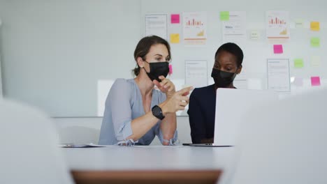 Two-diverse-female-colleagues-wearing-face-mask,-sitting-at-desk,-looking-at-laptop-and-talking