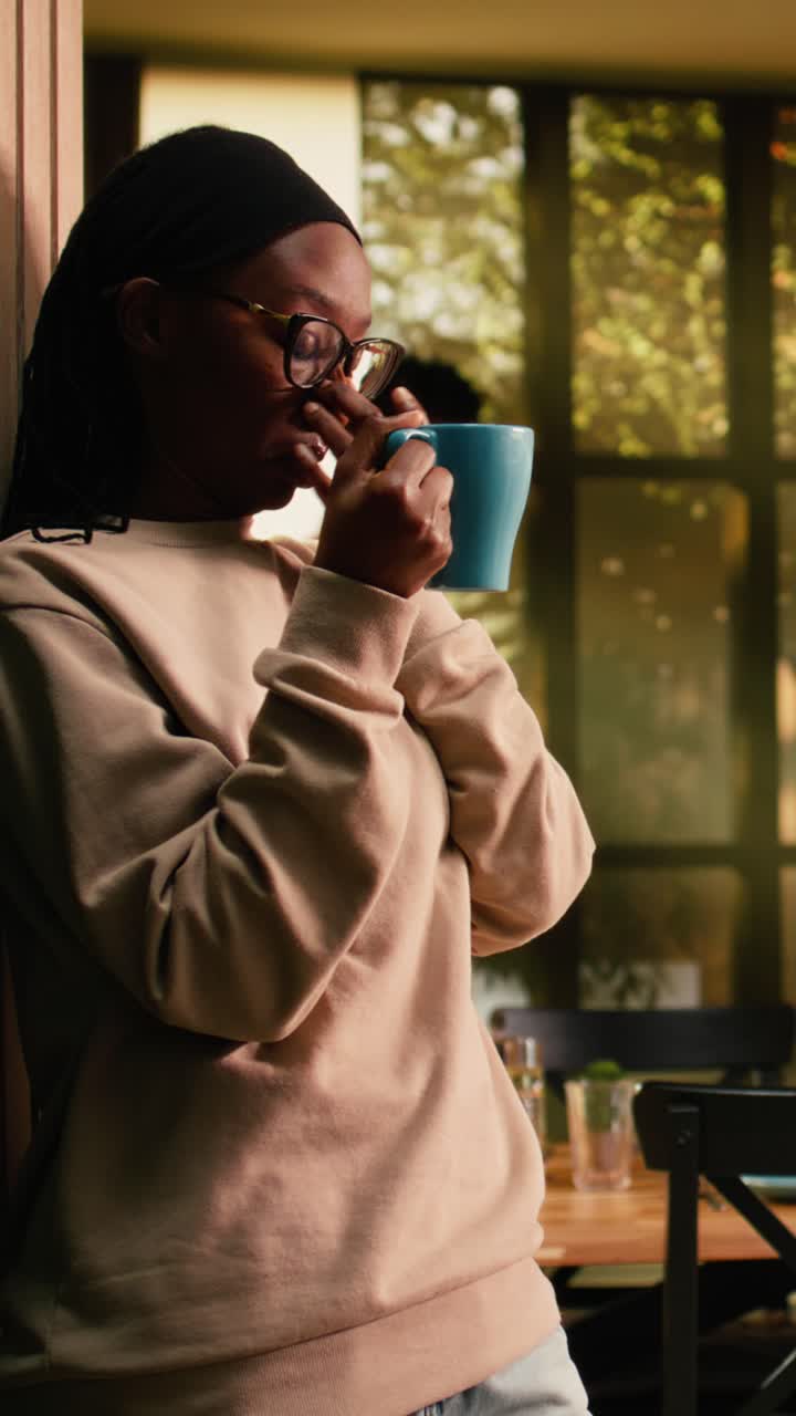 Video verticale ragazza afroamericana, che beve una tazza di caffè, che si rilassa in casa, che serve una bevanda