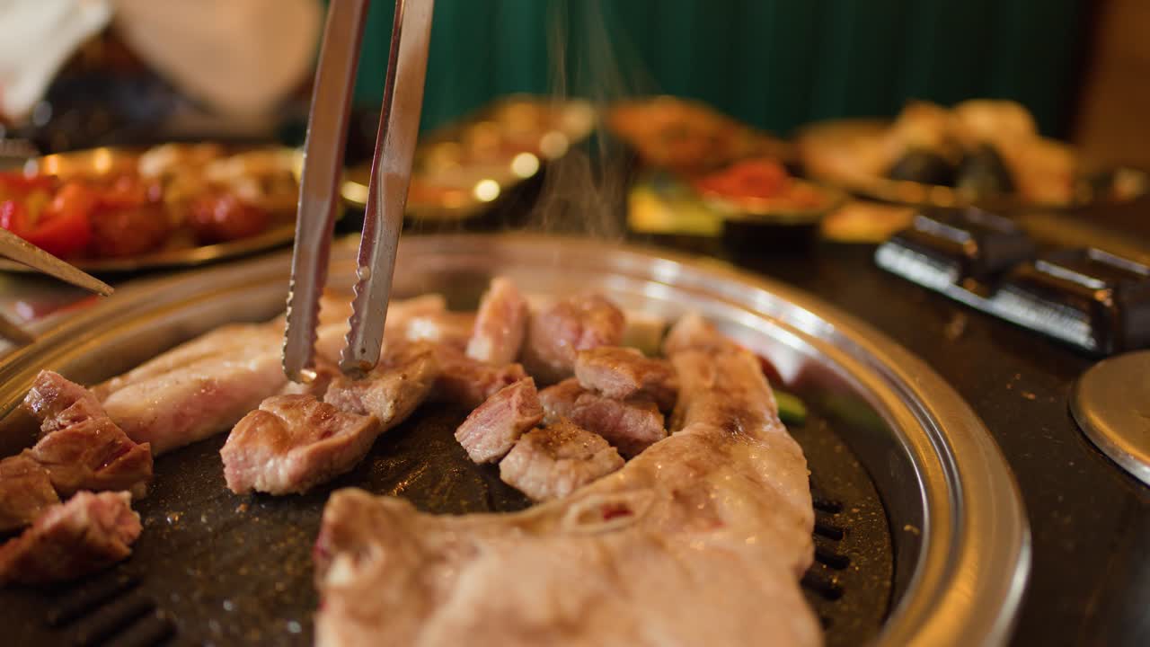 Penjepit memotong perut babi yang mendesis di atas panggangan meja, pencahayaan hangat, kedalaman dangkal, suasana restoran