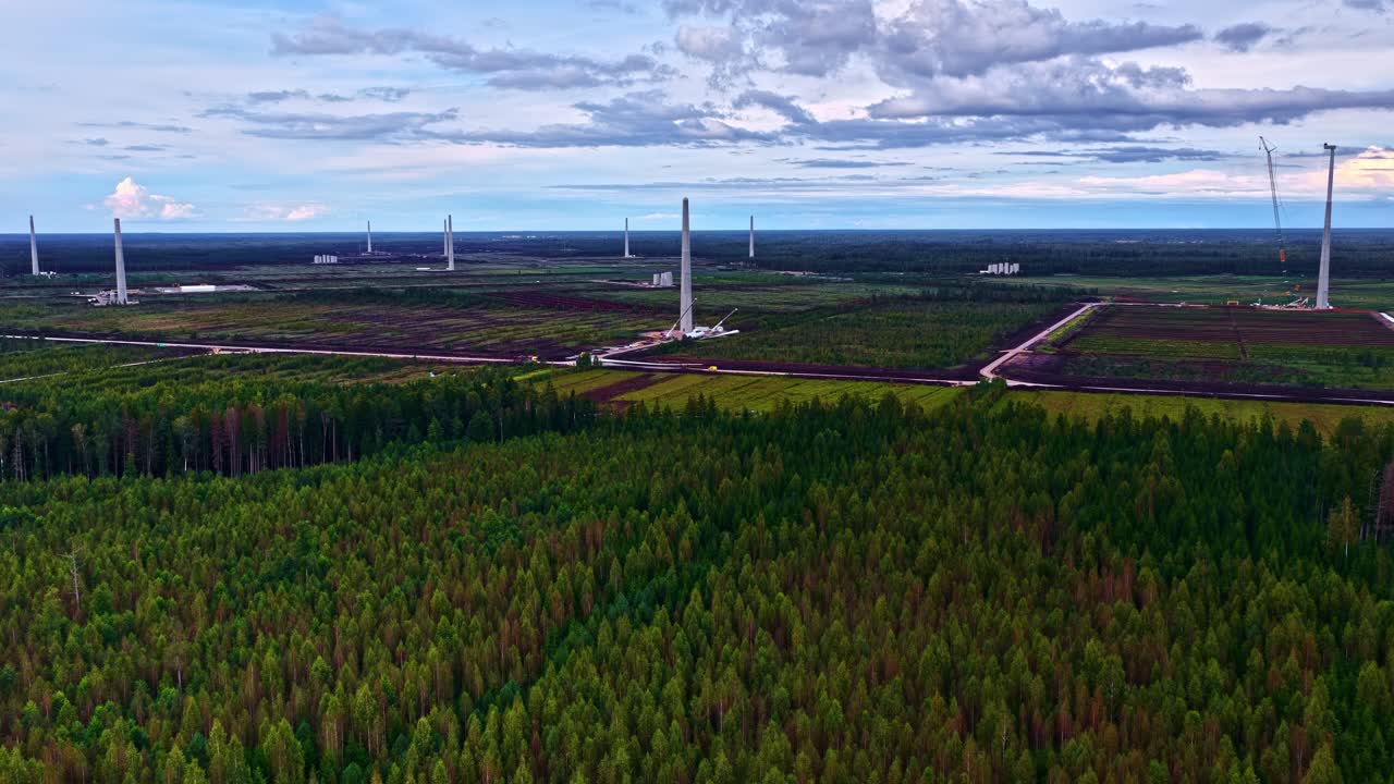 Several wind turbine towers are under assembly with cranes on cleared land amid surrounding forest