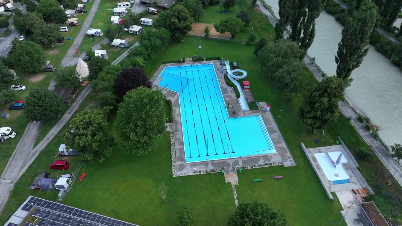 Luftfoto med udsigt over en campingplads' swimmingpool, beliggende langs en hurtigtstrømmende schweizisk flod.