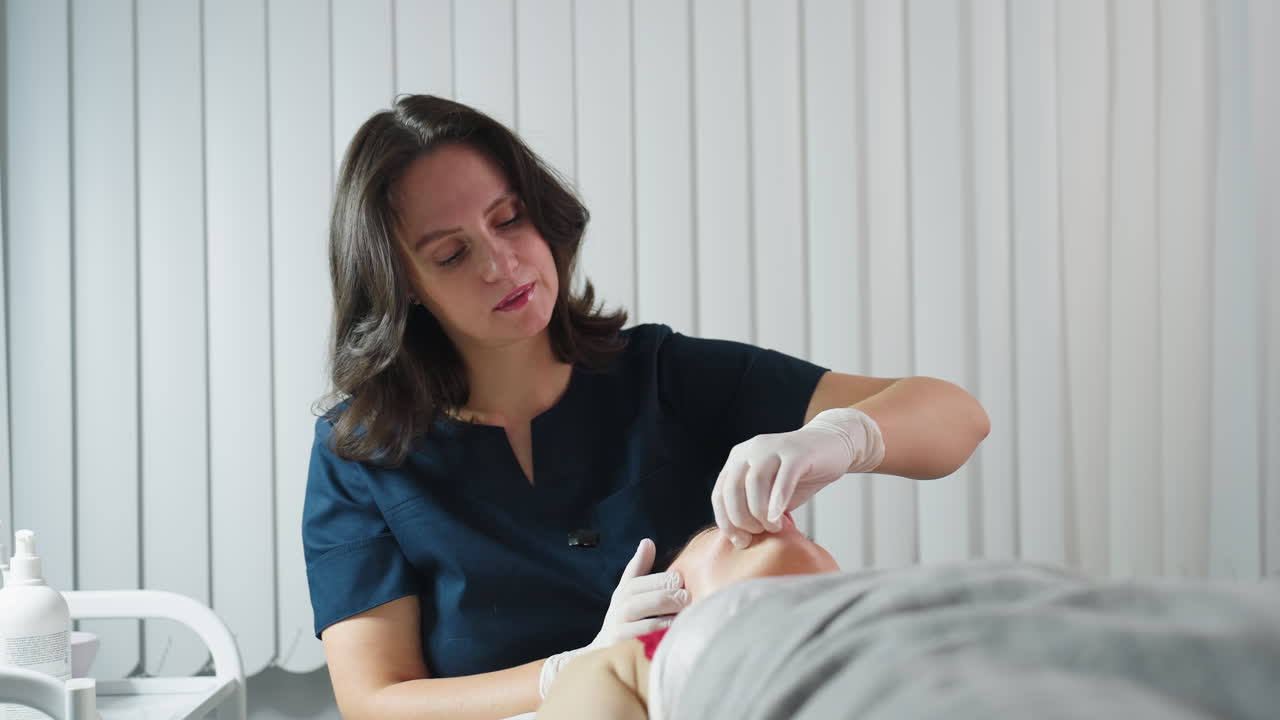 Close up of aesthetician wearing navy clinical uniform and safety gloves dipping fingernail inside client mouth while gently massaging jawline and side of face during session in clinic setting