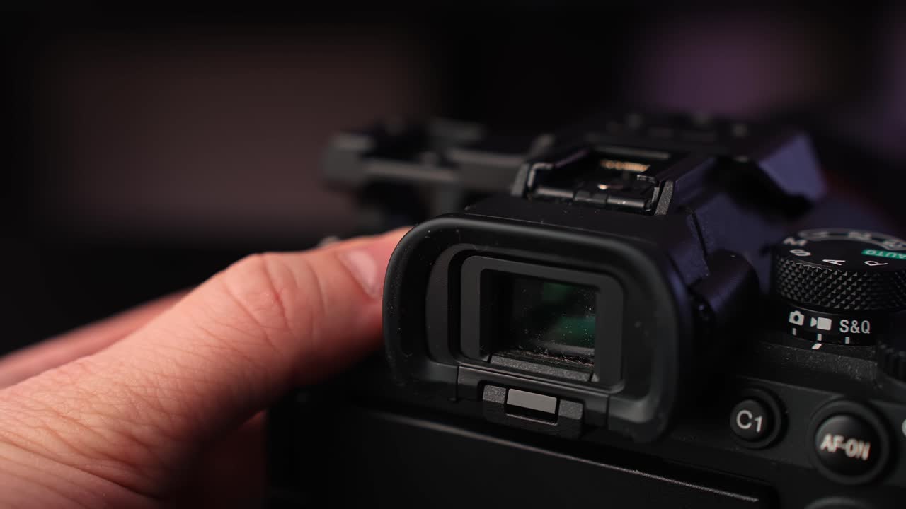 Hand grips mirrorless body while framing through the electronic viewfinder, showing top dials and controls in moody studio light with shallow depth for creator tech b-roll