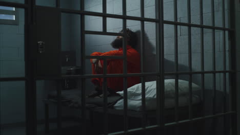 guilty criminal in orange uniform sits on the bed in prison cell