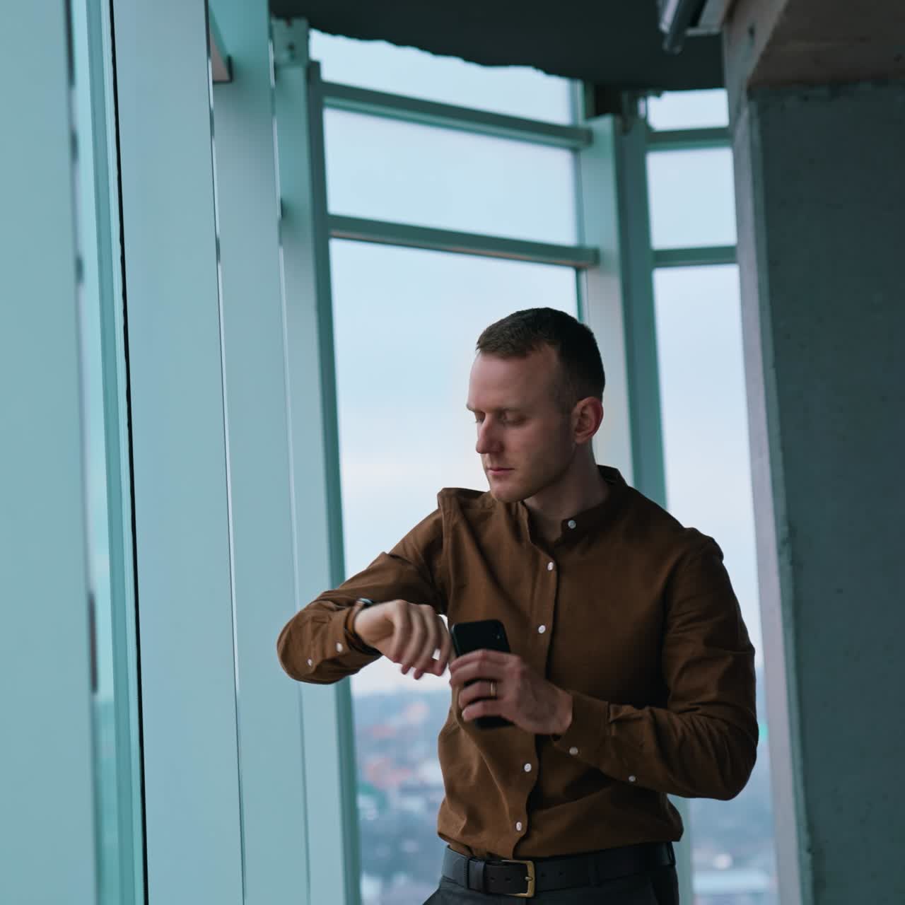 Serious businessman looking through the window. Thoughtful young man waiting for someone and looking on watch in modern office