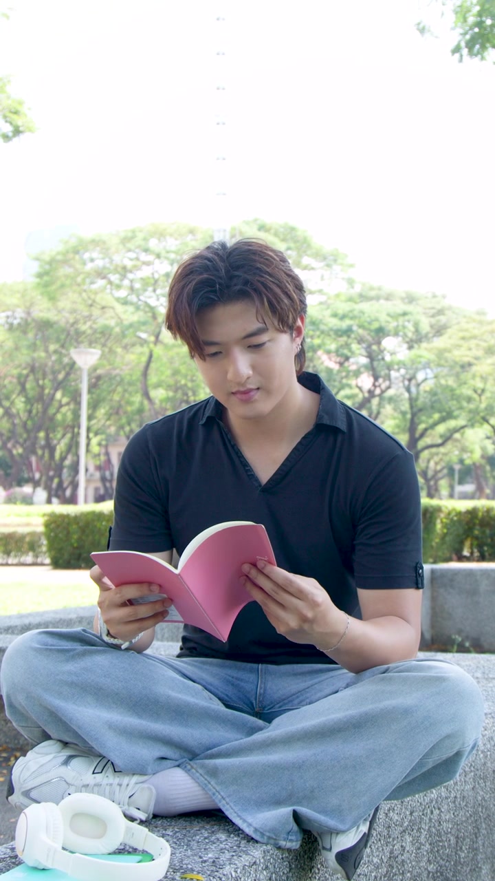 ชายหนุ่มชาวไทยนั่งขัดสมาธิในสวนสาธารณะที่มีแดดจ้า โดยตั้งใจอ่านหนังสือ