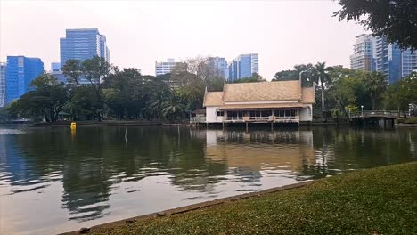 City-park-lake-with-fountain-in-the-foreground,-skyscrapers-in-the-background-at-dusk,-serene-mood,-Lumpini-Park-in-Bangkok,-Thailand