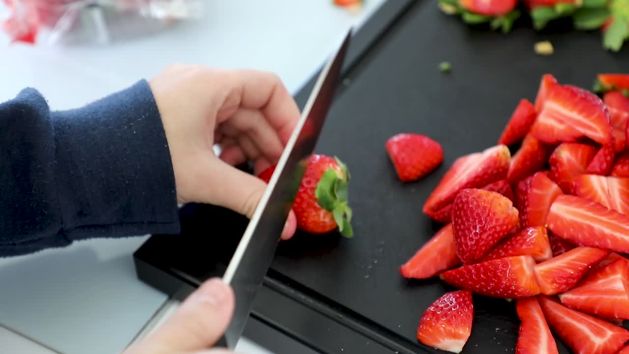 Close up of girl's hand removing strawberry leaves before slicing them in half with a knife. Pile of strawberry on black kitchen countertop