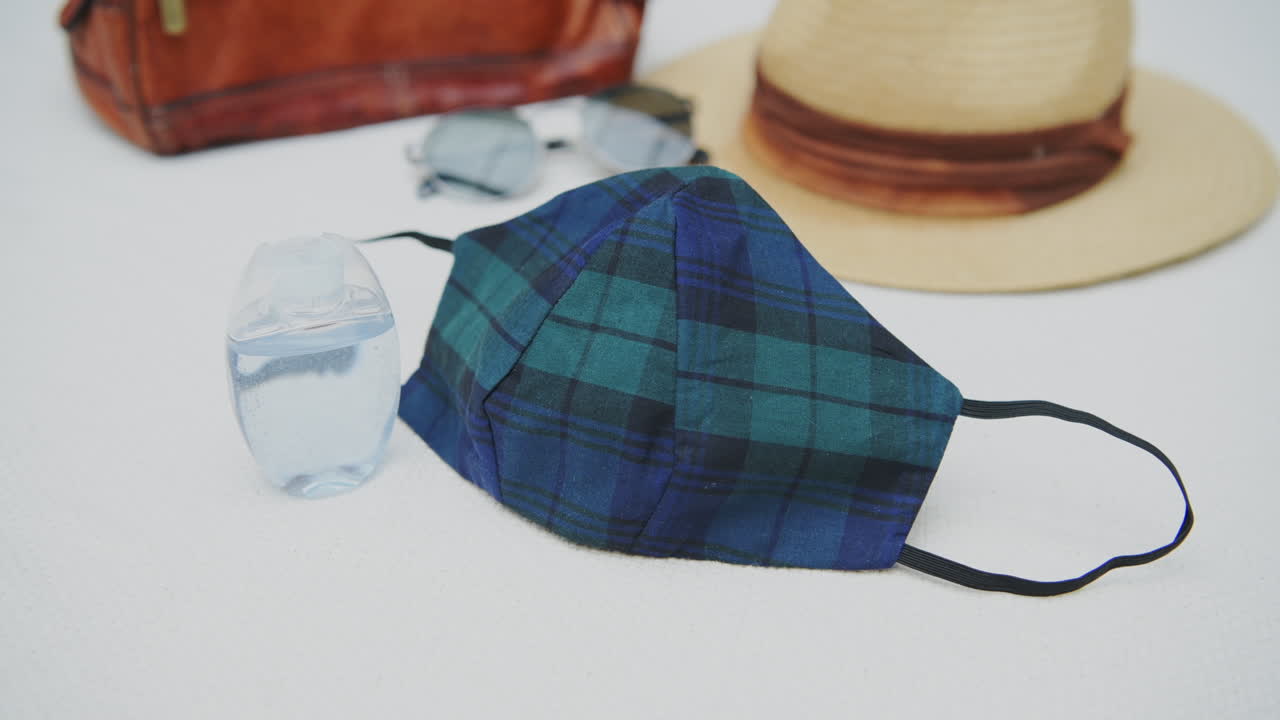Home made coronavirus face mask with plaid pattern, small bottle of hand sanitizer and travel items in background, flat lay, concept