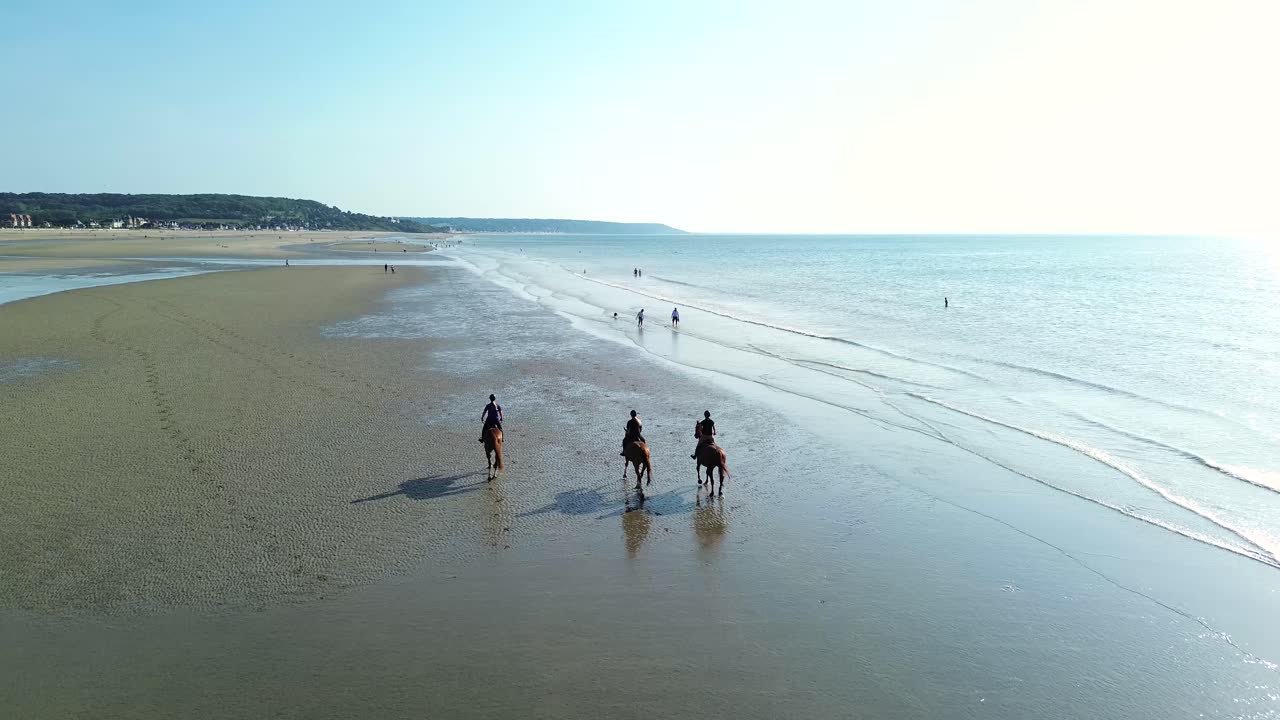 Drone shots of riders galloping at full speed across the reflective shoreline of Deauville beach. The sun glistens on the water as hooves kick up spray,